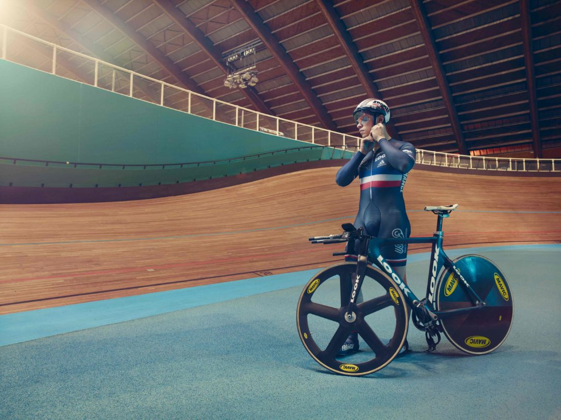Cyclist in a dark racing skinsuit stands beside a track bicycle inside a velodrome with a wooden banked track.