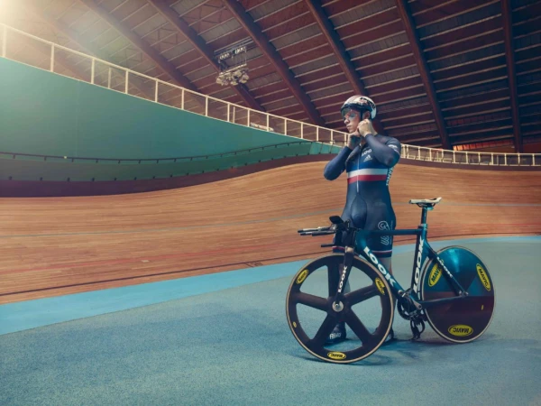 Cyclist in a dark racing skinsuit stands beside a track bicycle inside a velodrome with a wooden banked track.