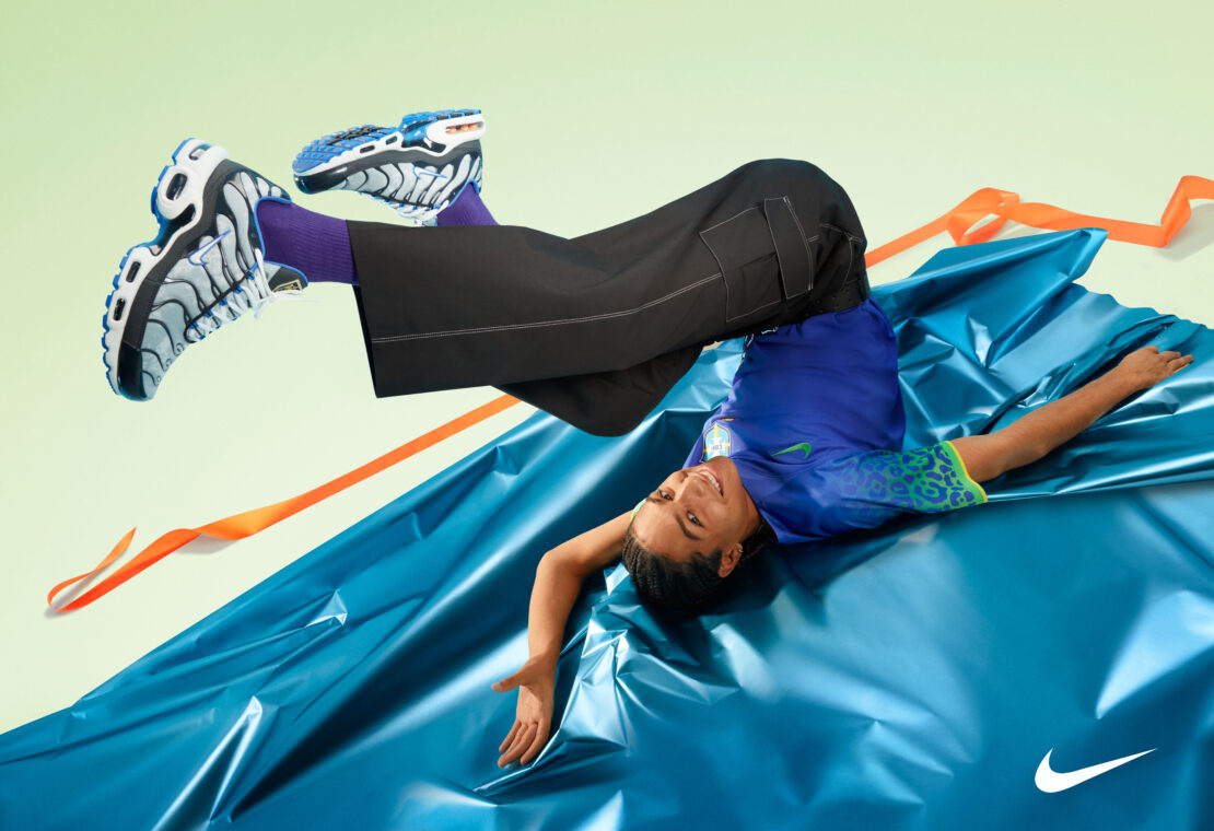 Athletic person in dynamic pose wearing sneakers and wide-leg pants on blue fabric, with Nike logo in view against pastel backdrop.