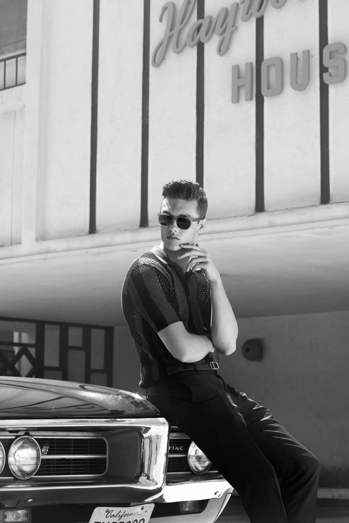 Man wearing sunglasses and a patterned shirt leaning on a classic car outside a building by Jean-Yves Lemoigne