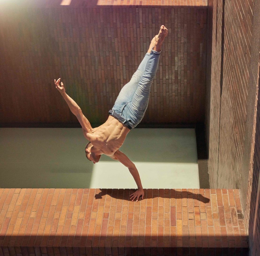 Shirtless man performing a one-arm handstand on a brick wall wearing blue jeans
