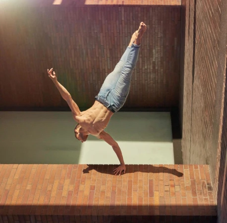 Shirtless man performing a one-arm handstand on a brick wall wearing blue jeans
