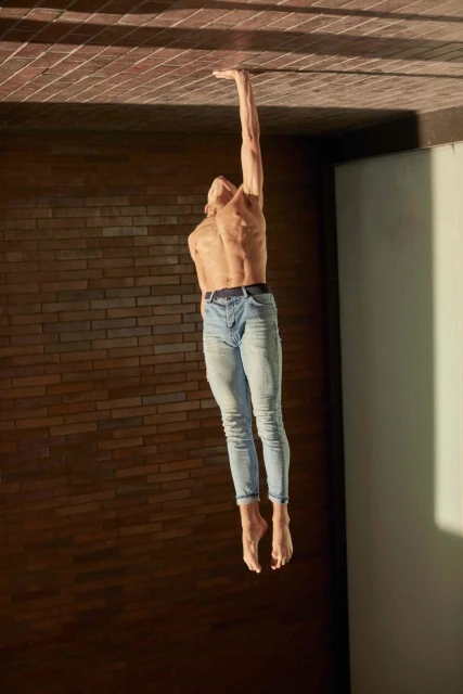Adult male in light blue jeans suspended, holding onto ceiling, in indoor setting with brick and light-colored walls, dramatic lighting.