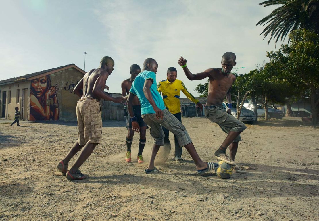 A group of young African men play barefoot soccer on a dusty pitch in a rural setting with a building and mural in the background.