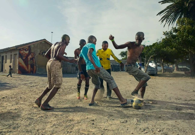 A group of young African men play barefoot soccer on a dusty pitch in a rural setting with a building and mural in the background.