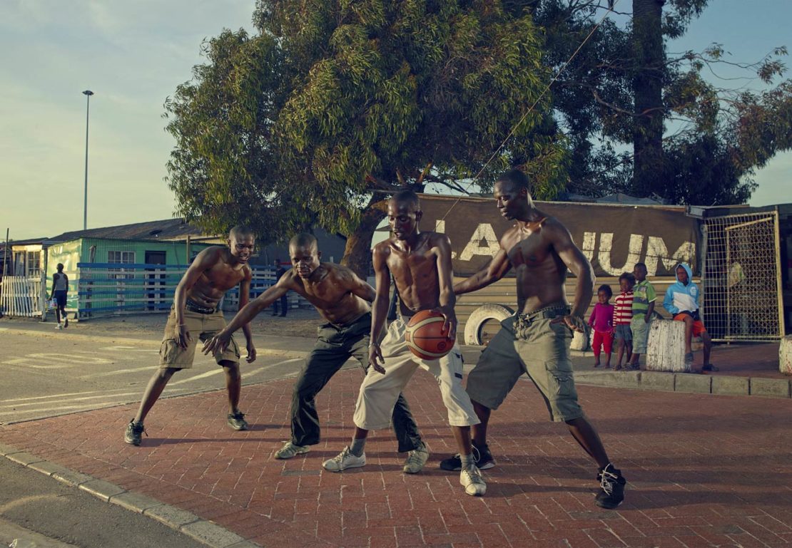 Four African adult males playing basketball on a red brick court, outdoor with a building and tree visible in the background.