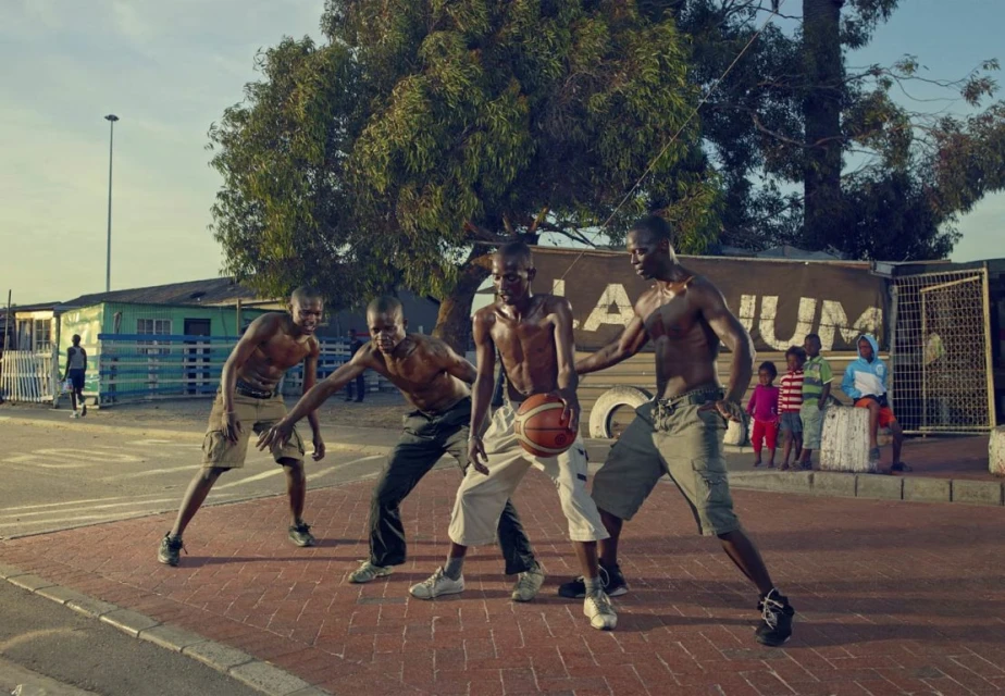 Four African adult males playing basketball on a red brick court, outdoor with a building and tree visible in the background.