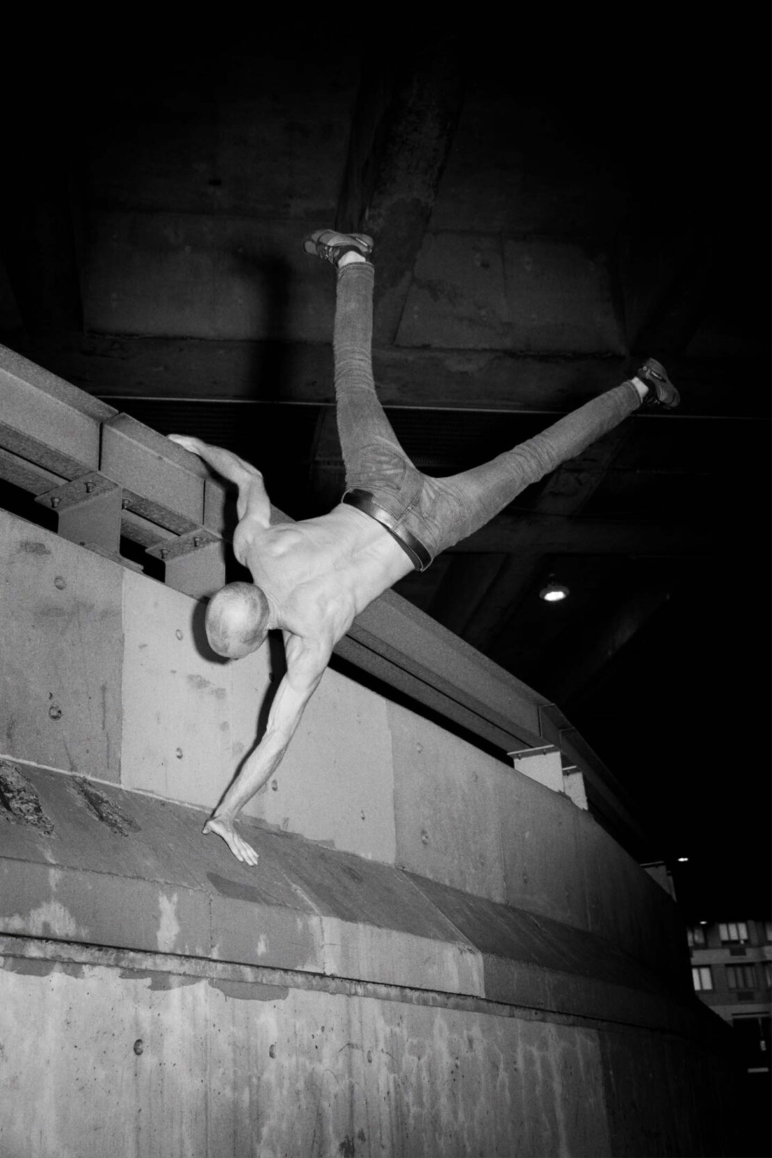 A shirtless man is doing a handstand on a concrete ledge, wearing jeans and balancing with his legs split in a dimly lit, urban environment.