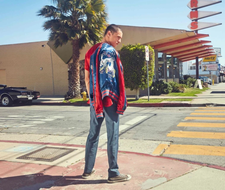 Man poses on crosswalk in jeans and red t-shirt, holding a jacket, with a black convertible car and diner in the background.