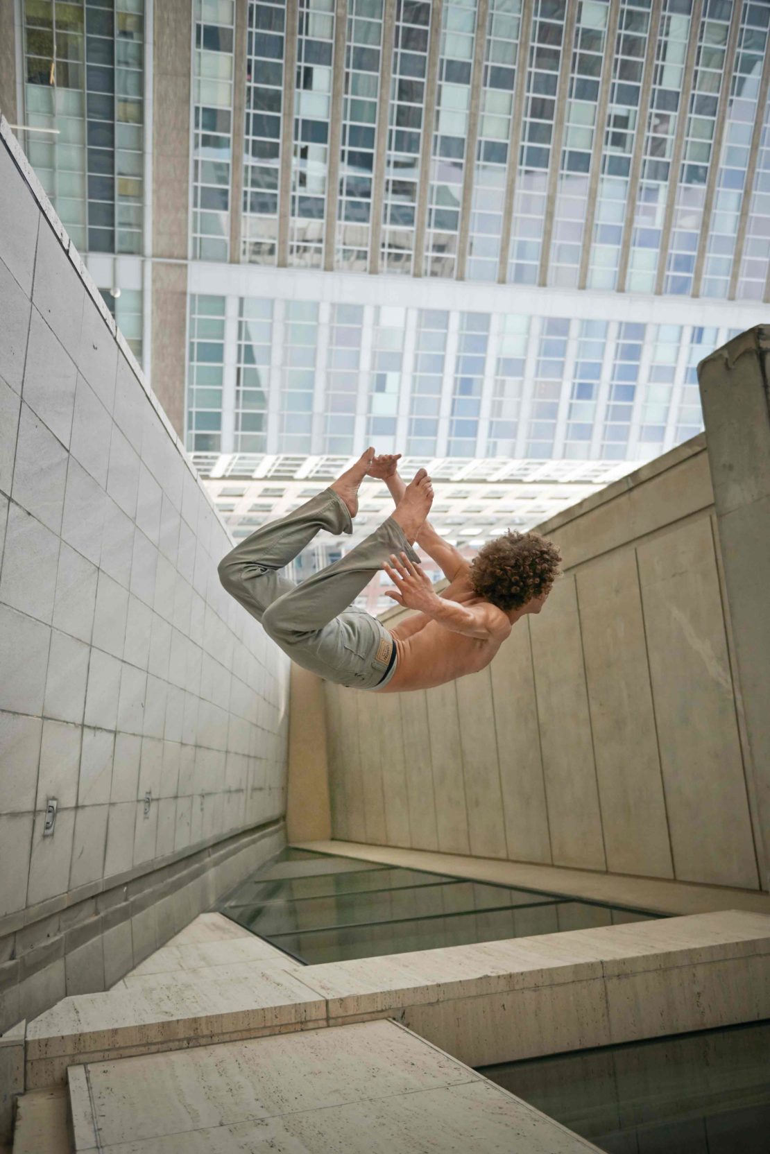 A man in gray pants performs acrobatics between concrete walls below a city building, captured from a low angle in a vertical shot.