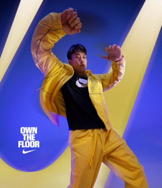 Asian man in yellow tracksuit dances against a blue backdrop with yellow accents, wearing black t-shirt with white logo.