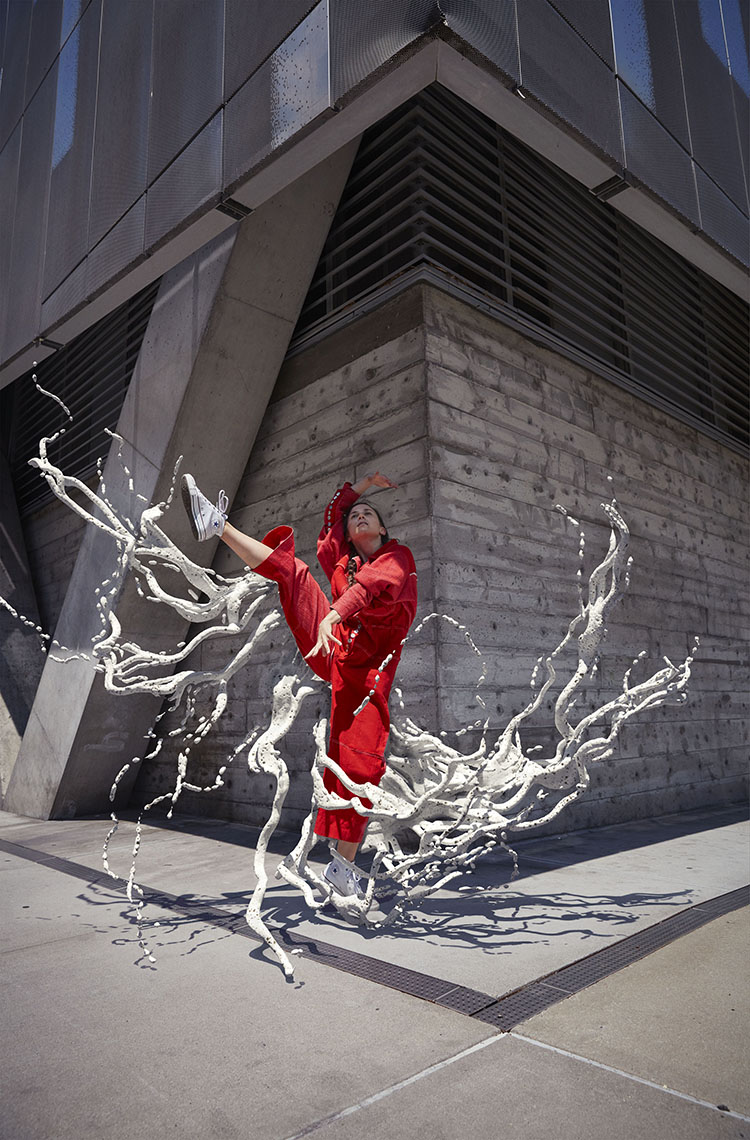 Adult female dancer in red outfit strikes a pose with raised leg, performing outdoors against a modern building and sculptural elements.