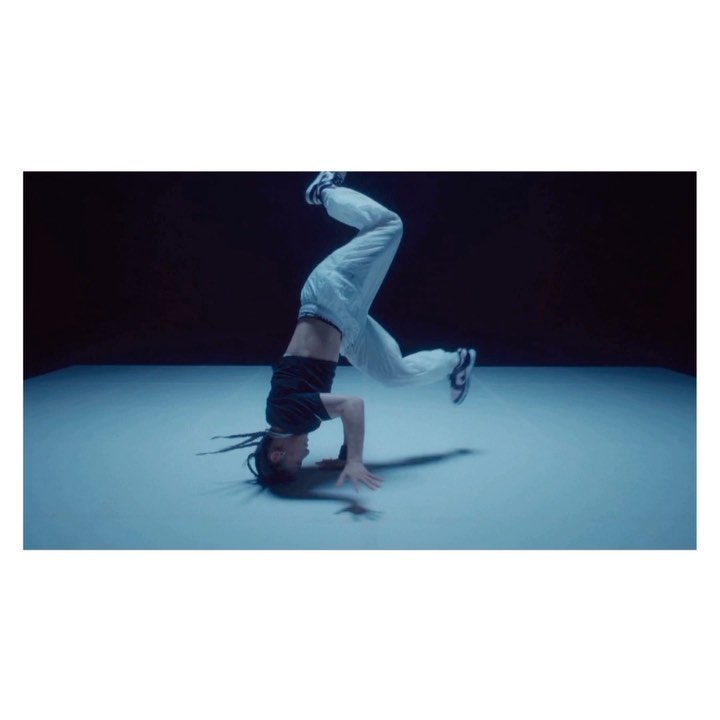 A person in a black crop top and white pants is doing a headstand on a light blue floor with a dark background, lit by a blue light.