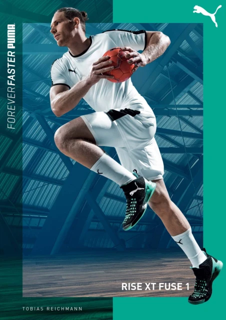 Male handball player in mid-air with ball wearing white uniform and black sneakers on court with teal and blue structural background.