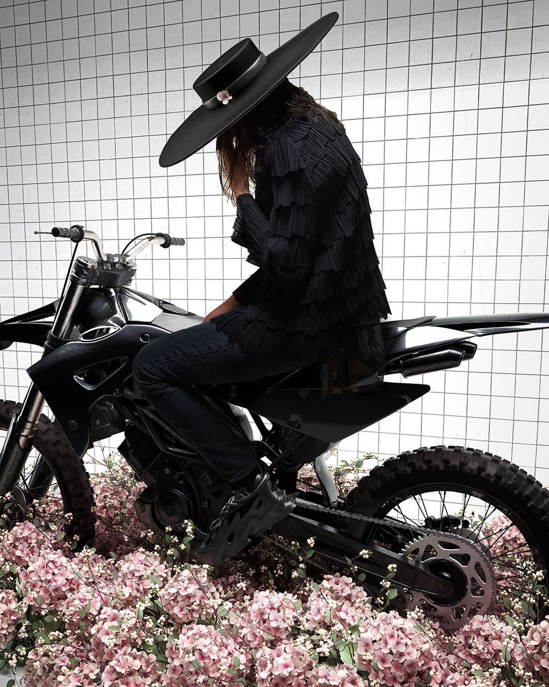 Person in black hat and fringed jacket sits on a dirt bike surrounded by pink flowers against a white grid backdrop in a studio setting.