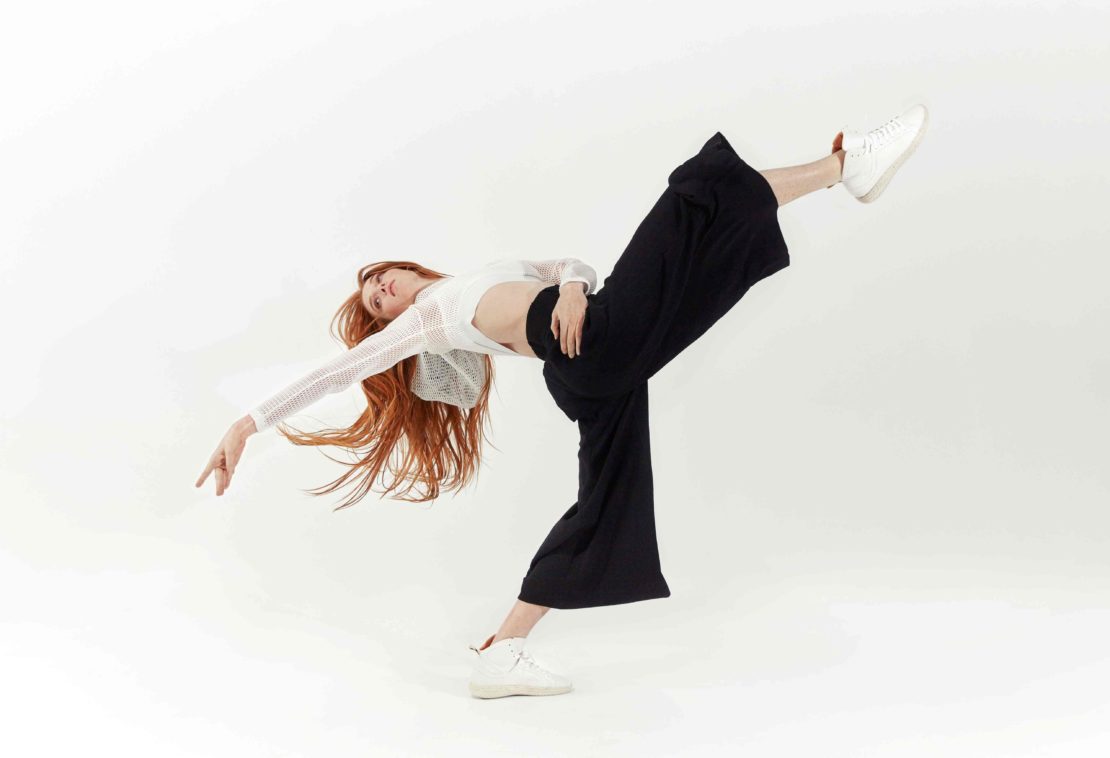 A woman with long red hair poses in a dynamic dance position wearing a white top, black pants, and sneakers against a white backdrop.