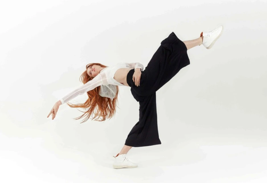 A woman with long red hair poses in a dynamic dance position wearing a white top, black pants, and sneakers against a white backdrop.