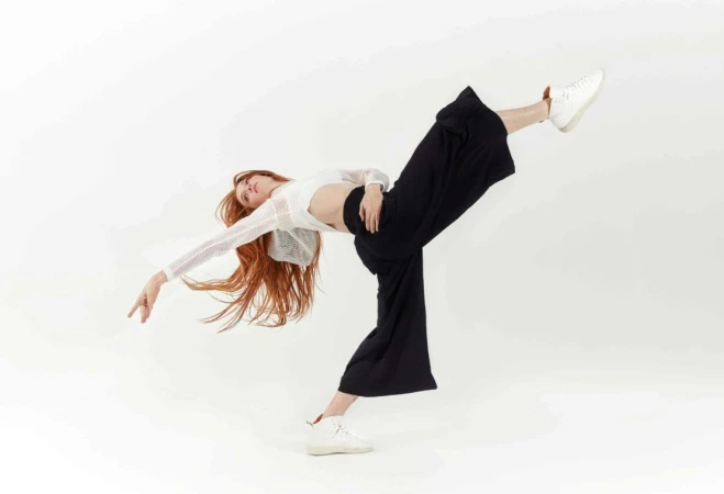 A woman with long red hair poses in a dynamic dance position wearing a white top, black pants, and sneakers against a white backdrop.