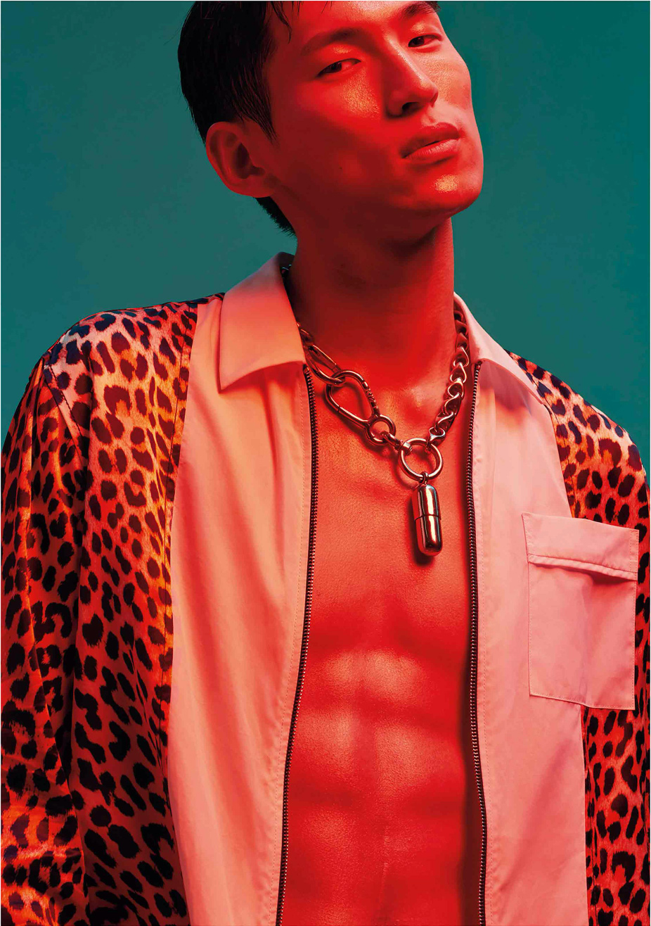 Portrait of an Asian male model in a vivid red light, wearing a leopard print shirt and silver chain necklace, against teal background.