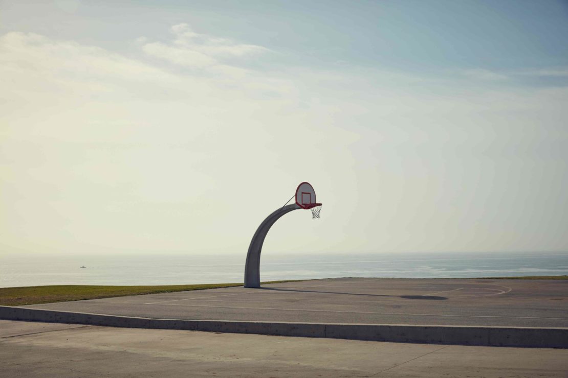 Outdoor basketball court featuring a concrete surface and a basketball hoop with an ocean background under a cloudy sky.