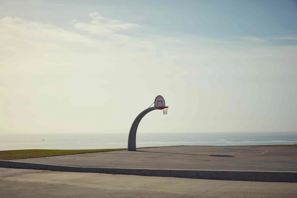 Outdoor basketball court featuring a concrete surface and a basketball hoop with an ocean background under a cloudy sky.