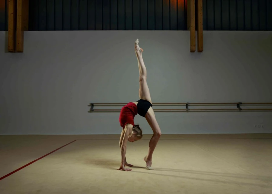 Female gymnast in red top and black shorts performing a backbend with one leg extended vertically in a gymnasium with beige floors.