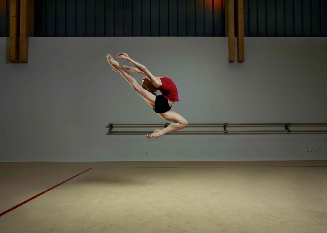 Female gymnast in mid-air leap, arching her back in an indoor practice space with a wooden bar and grey walls.