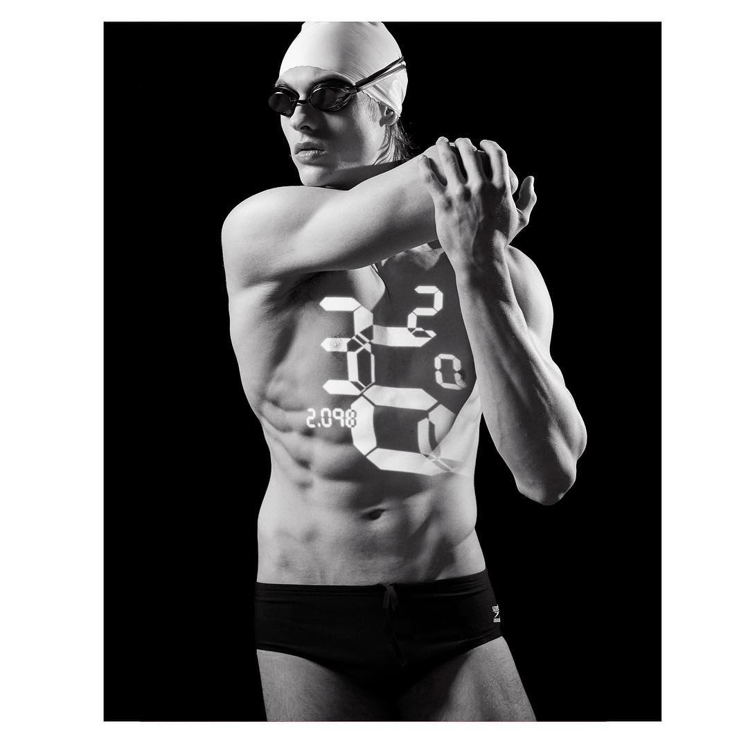 Monochrome image of a swimmer with a digital timer graphic overlay, wearing goggles and cap, in a dark studio setting.