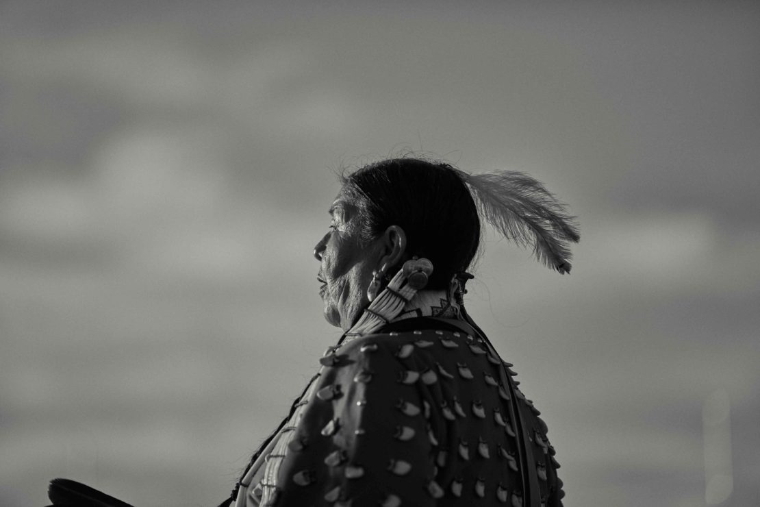 Profile of an Indigenous woman with a feather in her hair, wearing traditional dress, set against a soft, monochromatic sky background.
