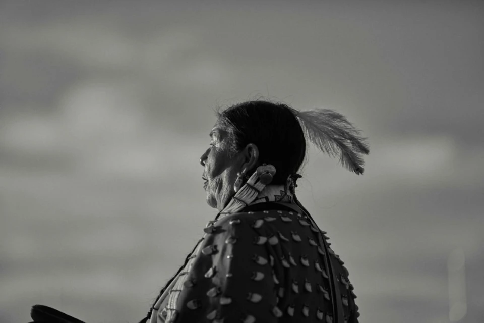 Profile of an Indigenous woman with a feather in her hair, wearing traditional dress, set against a soft, monochromatic sky background.