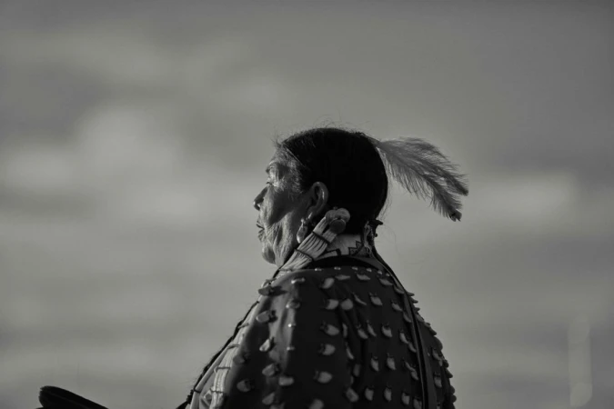 Profile of an Indigenous woman with a feather in her hair, wearing traditional dress, set against a soft, monochromatic sky background.