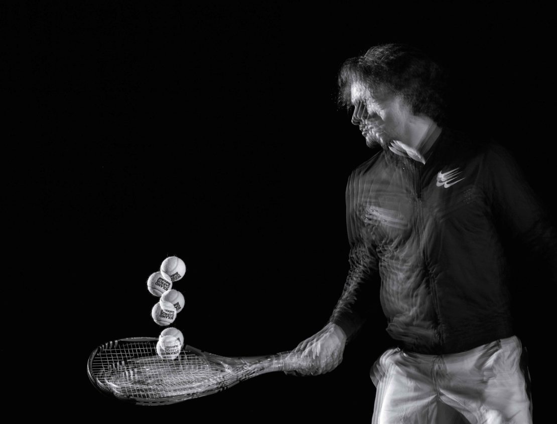 Action shot of a male athlete hitting tennis balls with a racket on a black background. Motion blur shows the speed and movement.