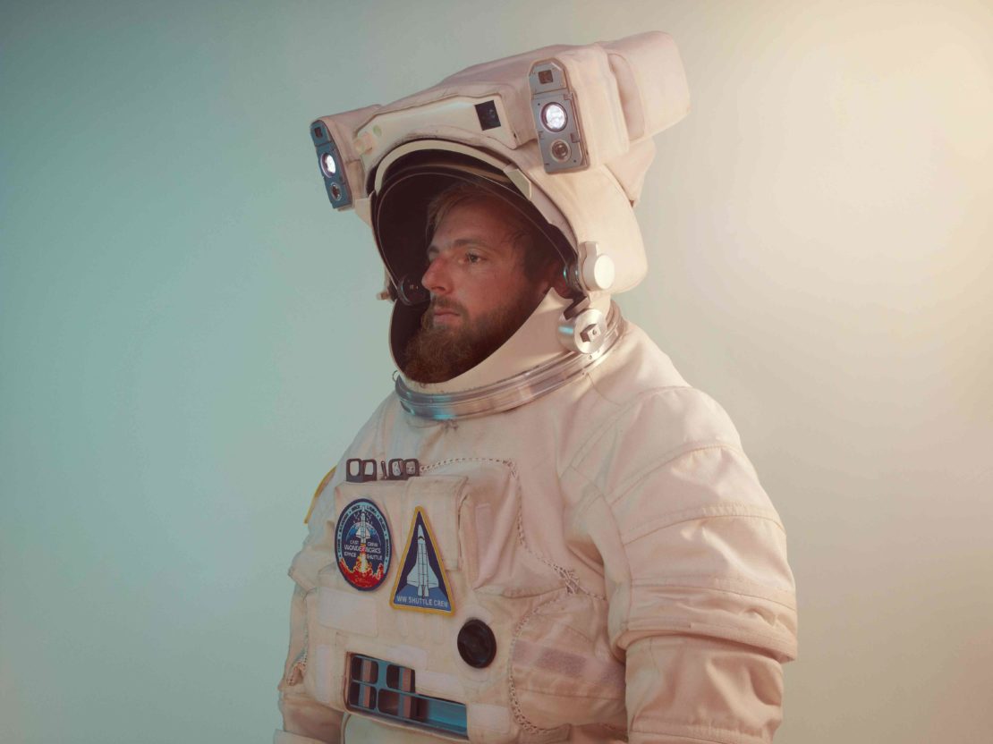 Side view of a bearded adult male in an astronaut spacesuit with patches against a light-blue background in a studio shot.