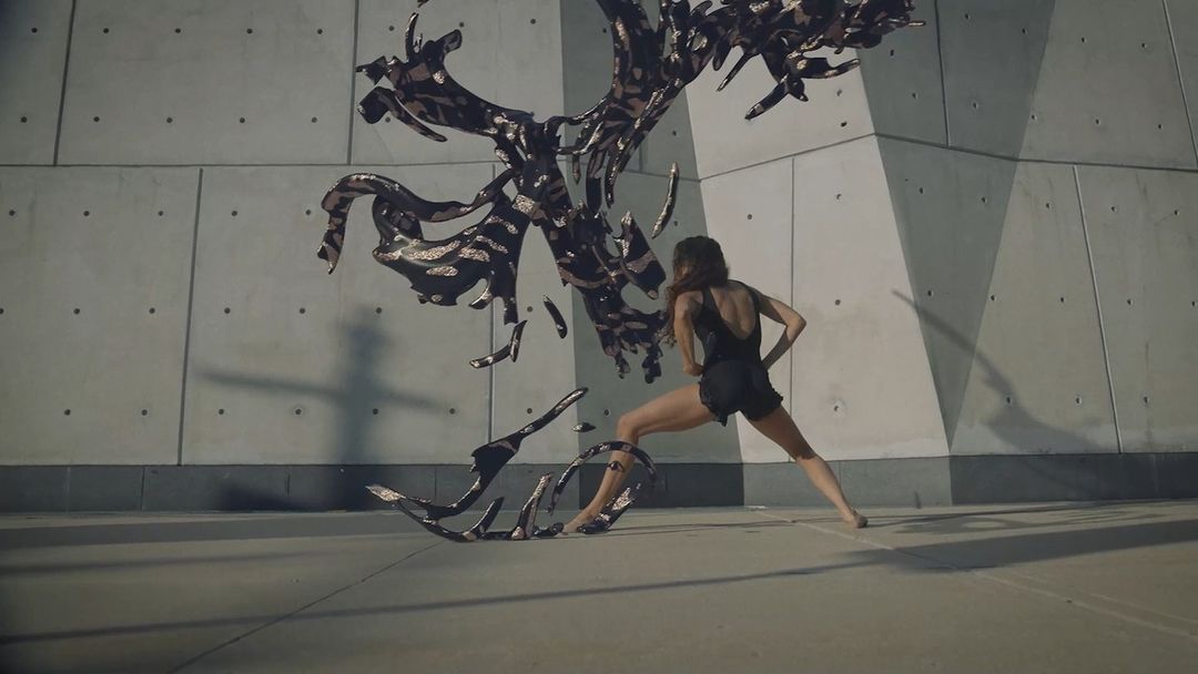 An adult female performs modern dance in front of a concrete wall with a fluid, dark abstract sculptural element on a concrete floor.