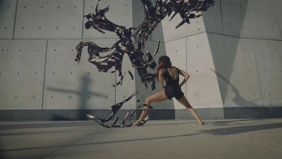 An adult female performs modern dance in front of a concrete wall with a fluid, dark abstract sculptural element on a concrete floor.