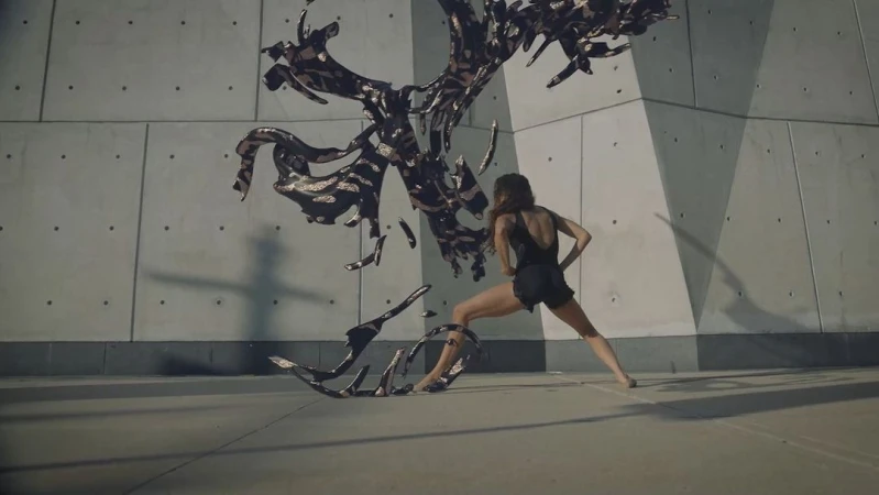 An adult female performs modern dance in front of a concrete wall with a fluid, dark abstract sculptural element on a concrete floor.