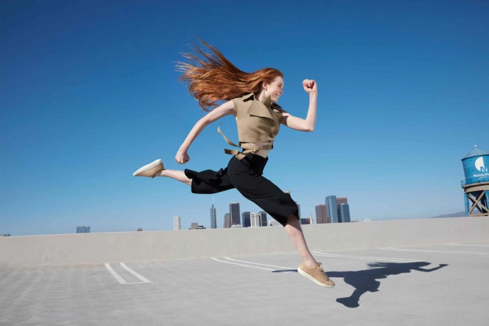 Woman with long red hair leaps in the air on a city rooftop, wearing a tan sleeveless top and black pants against a clear blue sky.