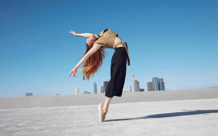 Red-haired female dancer leaping and arching backward on a rooftop against a clear blue sky