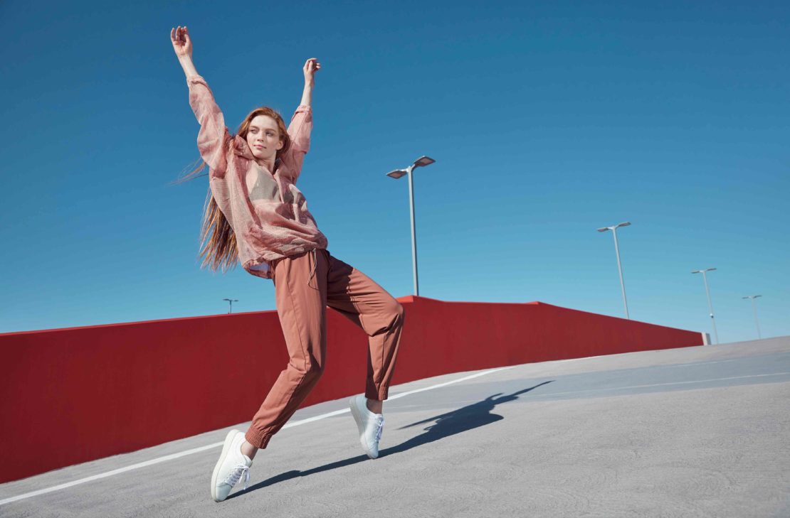 Redheaded woman dancing outdoors in pink translucent blouse and terracotta trousers against a clear blue sky