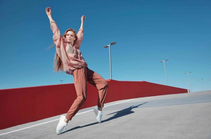 Redheaded woman dancing outdoors in pink translucent blouse and terracotta trousers against a clear blue sky