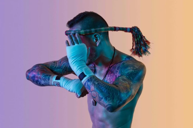 Shirtless man with tattoos wearing a headdress and hand wraps, posing against a gradient background of orange, pink and blue.