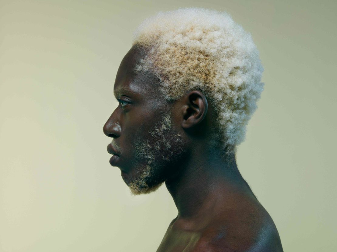 Profile view of a dark-skinned man with blond afro hair against a pale yellow background, showing his face, neck and bare shoulder.