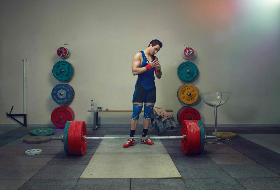 Weightlifter preparing at indoor gym. Man athlete wraps wrists near barbell and weights. Fitness training with red and blue sportswear.
