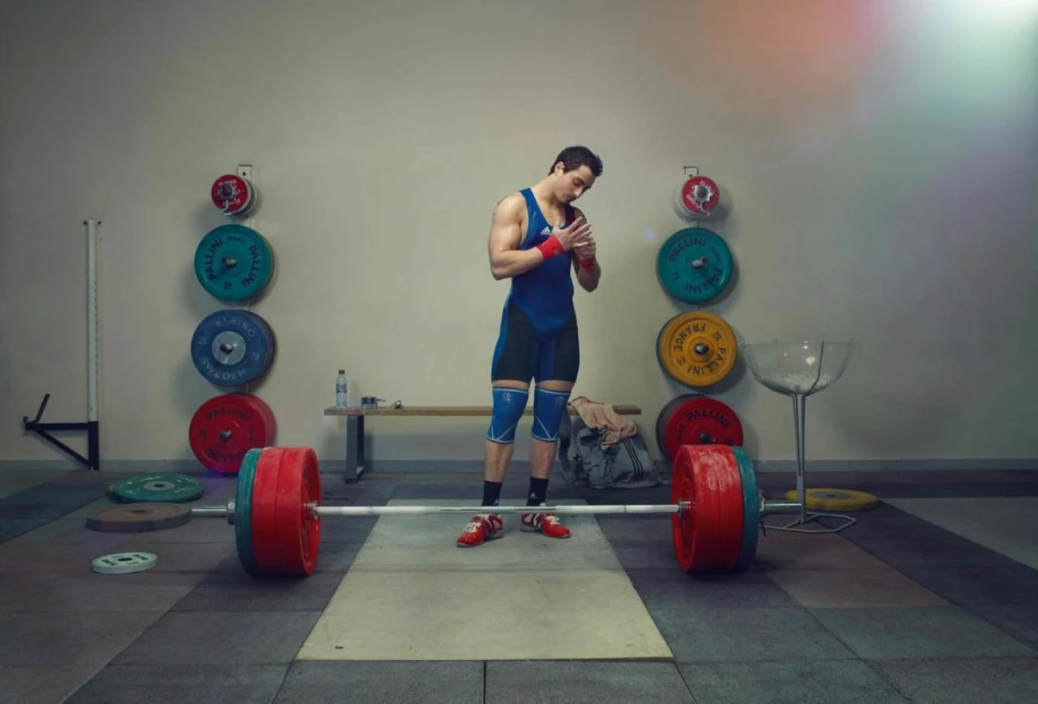 Weightlifter preparing at indoor gym. Man athlete wraps wrists near barbell and weights. Fitness training with red and blue sportswear.