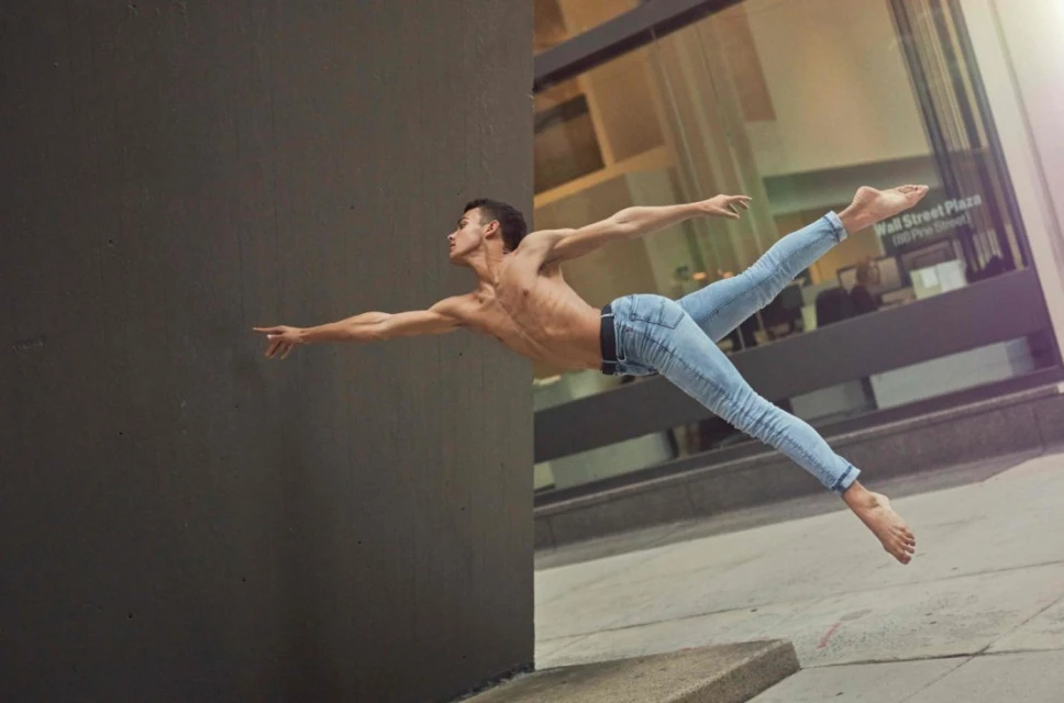 A shirtless man wearing jeans is leaping outdoors with his arms extended, appearing to fly between a concrete wall and a building.