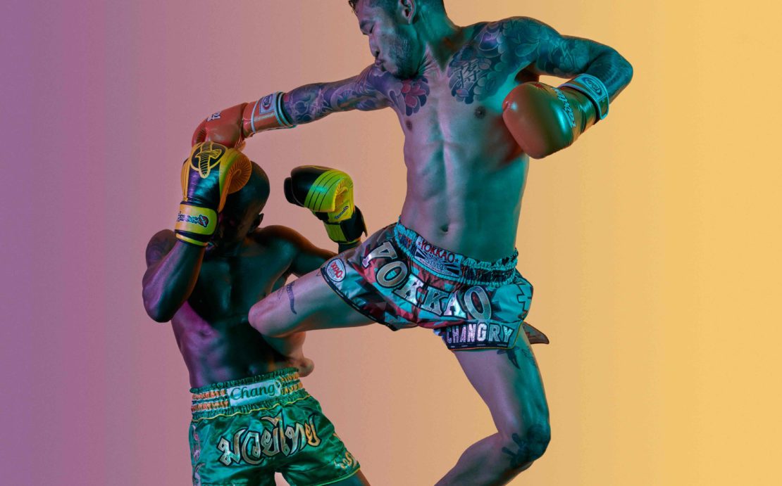 Two adult males engaged in a Muay Thai match, one striking while airborne, wearing boxing gloves and traditional shorts against a gradient backdrop.