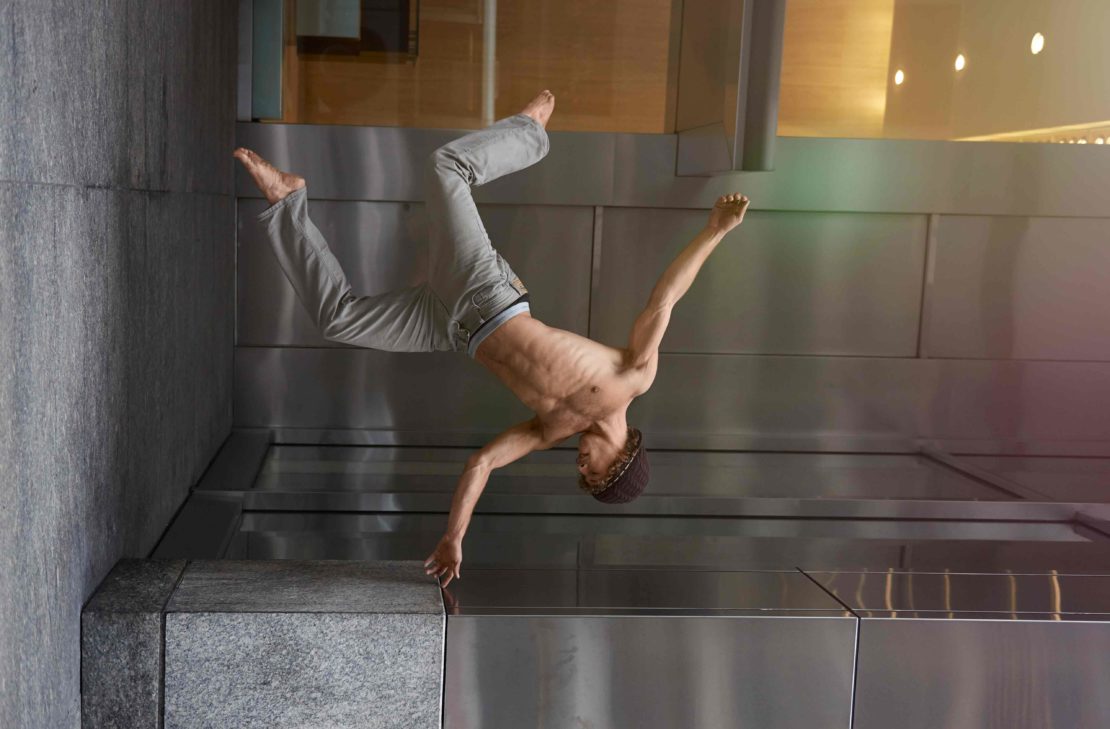 Athletic man performs a one-handed handstand wearing casual pants and a beanie, posing against a textured silver wall indoors with overhead lighting.