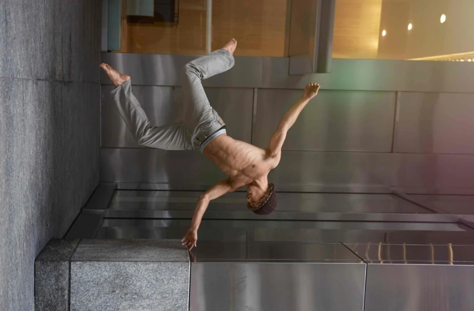Athletic man performs a one-handed handstand wearing casual pants and a beanie, posing against a textured silver wall indoors with overhead lighting.