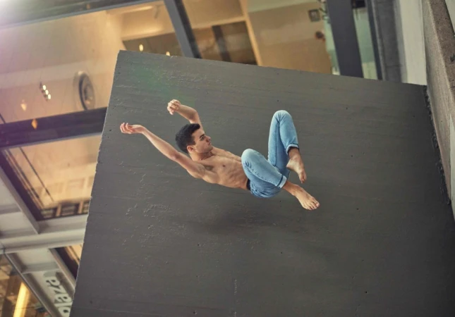 Shirtless man in blue jeans mid-air tumbling alongside a large concrete wall with modern architecture visible