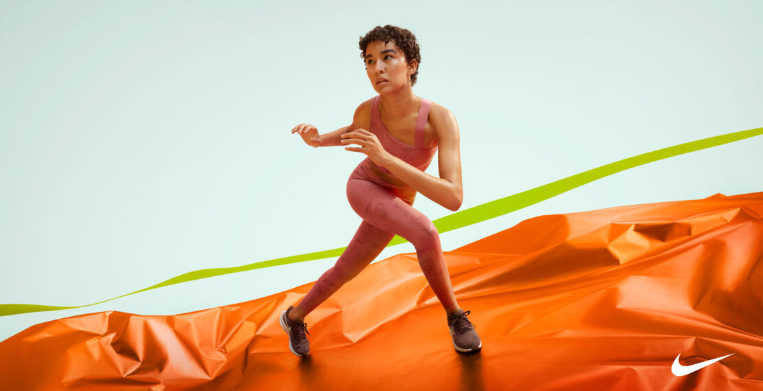 Athletic woman in pink sportswear lunging against a backdrop of shiny orange fabric and a light blue wall with a lime-green stripe.